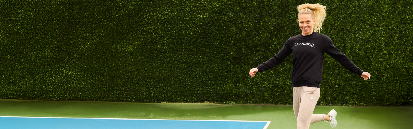 Model jumping up on pickleball court wearing the black 'play nicely' crew neck pull-over sweatshirt and beige super soft joggers